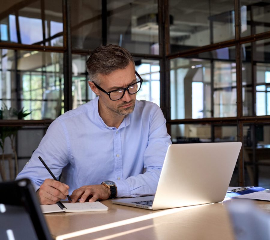 Mature business man executive manager looking at laptop computer watching online webinar training or having virtual meeting video conference taking notes, doing market research working in office.
